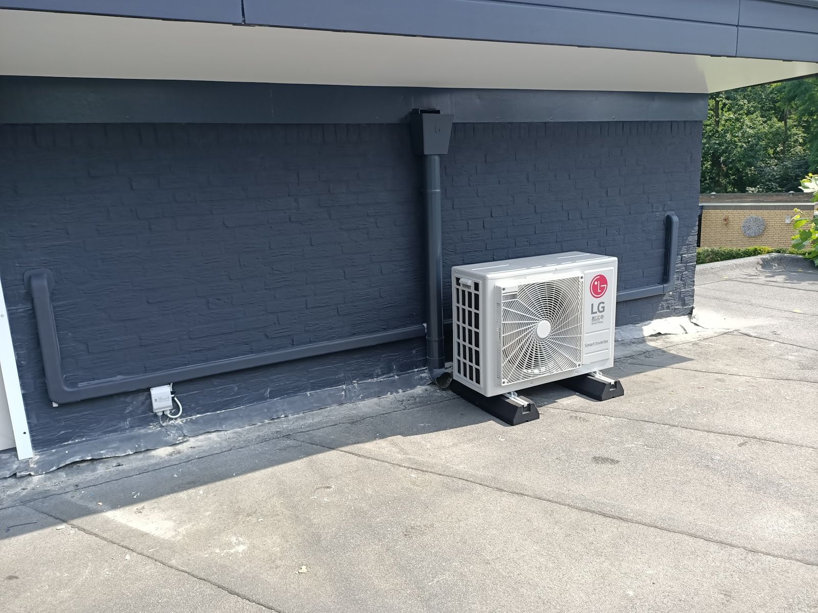 LG-buitenunit tegen een donkergrijze gevel, de leidingen in een geverfde goot