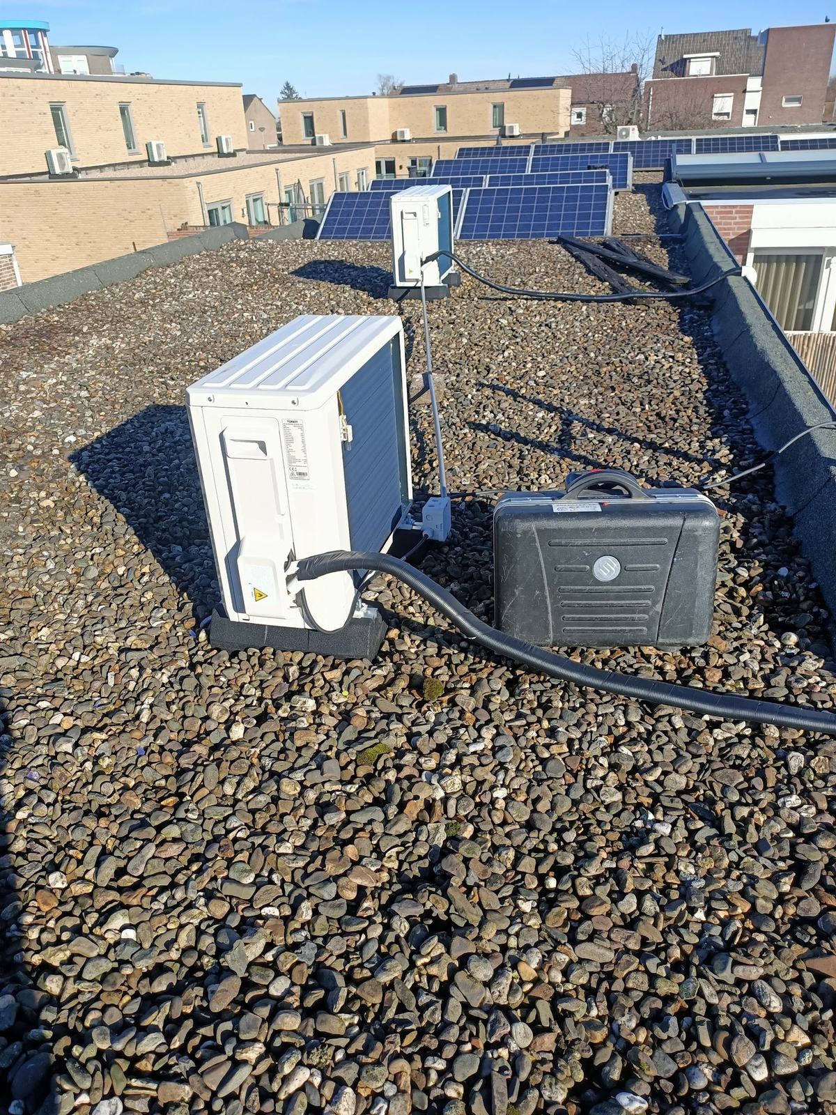 Buitenunit op een grindplat dak naast zonnepanelen, met een zwarte gereedschapskoffer ernaast