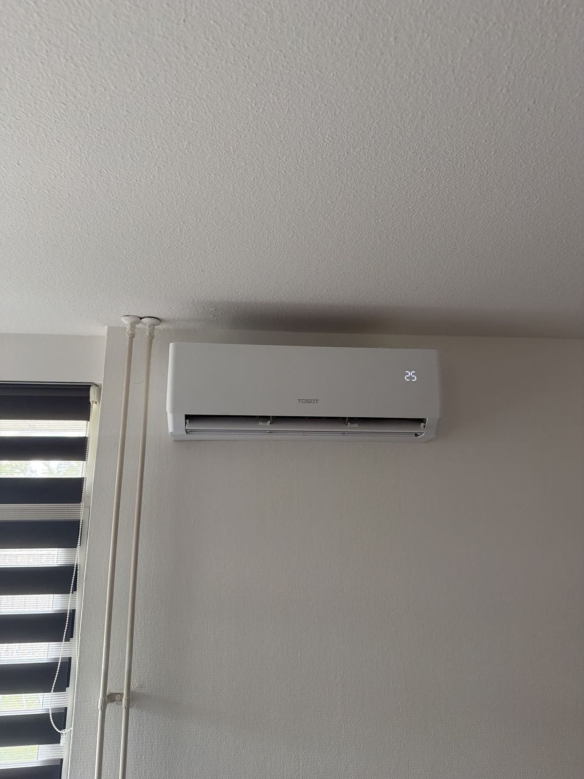 Witte binnenunit hoog aan de muur naast een raam met een rolgordijn